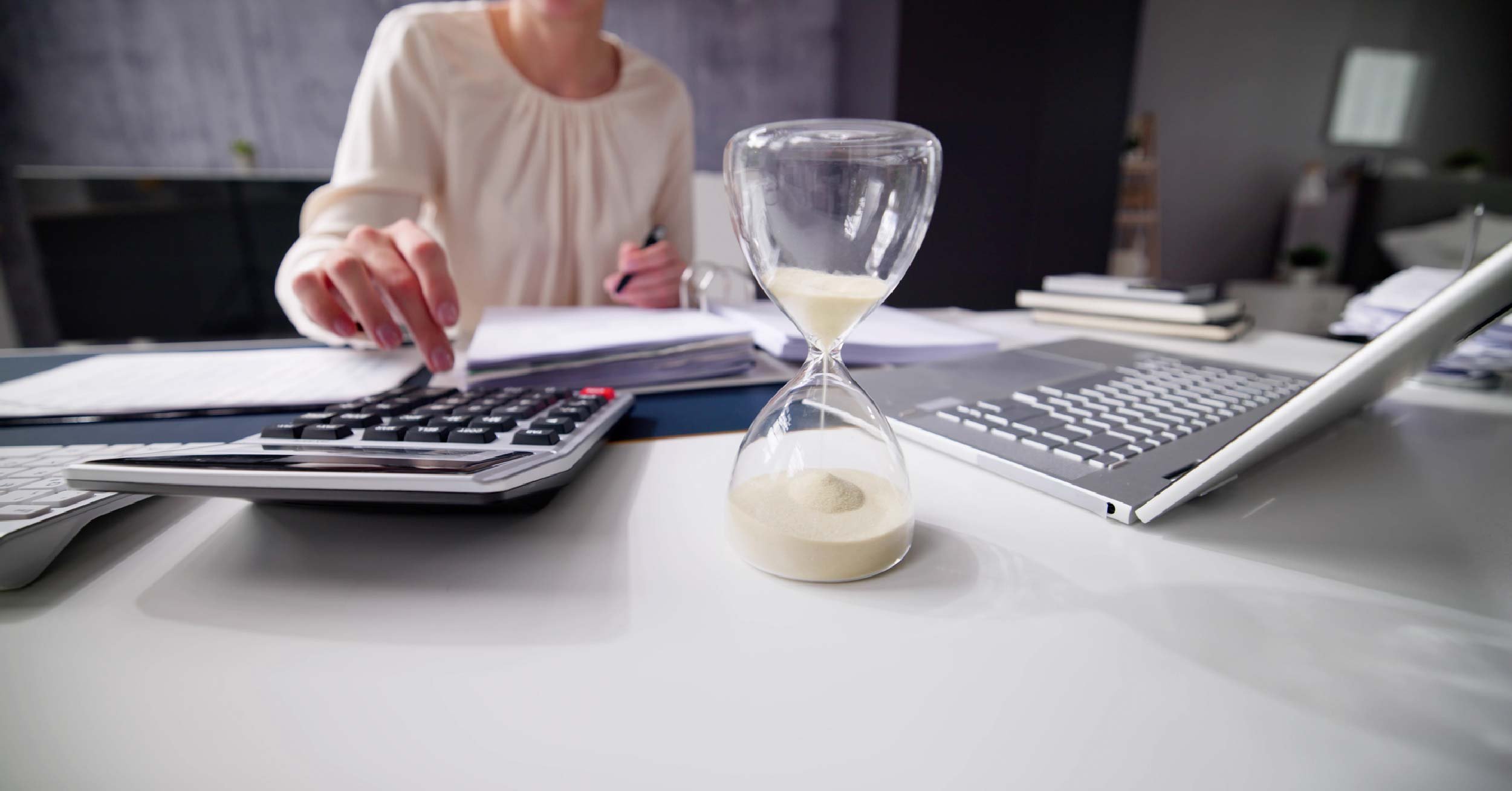 A female executive doing research on her laptop and making quick calculations as an hourglass with white sand slowly runs out.